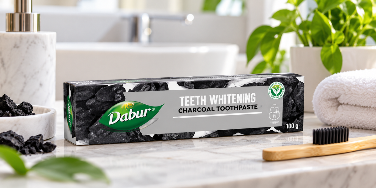 Dabur Charcoal Toothpaste 100g box displayed on a light marble bathroom countertop beside a bamboo toothbrush with dark bristles, pieces of charcoal, a folded white towel, and a green plant. The bright, modern bathroom setting highlights the toothpaste packaging for an online personal care product listing.
