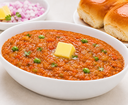 A white oval bowl filled with a thick orange-red curry or chutney, topped with a square of butter and small green herb pieces, with blurred bread rolls and a small bowl of chopped red onion in the background.