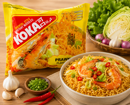 A bowl of Koka Prit Prawns Flavor instant noodles served with shrimp, green onions, red chili peppers, and peas, next to its yellow packaging bag on a wooden table with fresh vegetables.