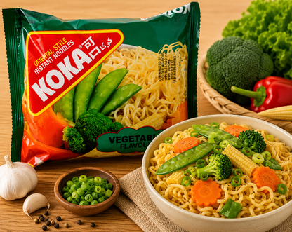 A package of Koka Veggie Flavor Instant Noodles stands next to a bowl of prepared noodles mixed with broccoli, snap peas, carrots, and corn, with garlic, peppercorns, and fresh vegetables on a wooden table.