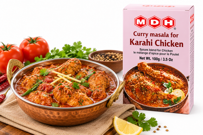 MDH Karahi Chicken Masala 100g spice blend packet displayed beside a traditional Indian-style chicken karahi curry in a copper serving bowl. The rich red-orange chicken curry is garnished with fresh cilantro and ginger strips, with ripe tomatoes, green chilies, lemon, and whole spices arranged nearby. The pink MDH packaging clearly displays the product name and illustrates the prepared chicken dish, making it suitable for showcasing an Indian cooking spice mix.