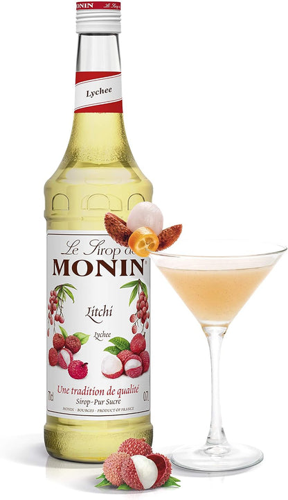 A bottle of Monin Lychee syrup next to a filled martini glass on a white background, with lychee fruits and flowers as decorative props.