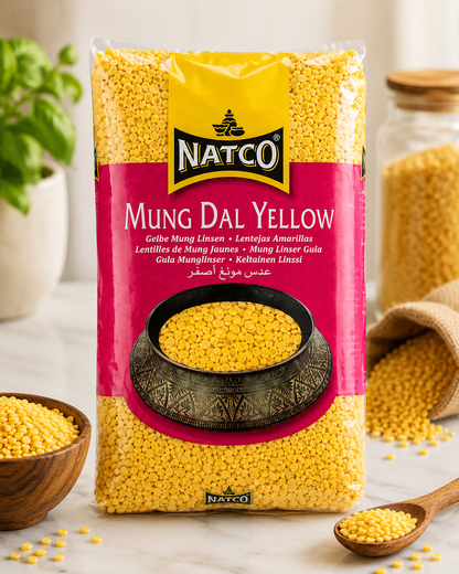 A bag of NATCO Mung Dal Yellow (yellow lentil) standing upright on a light marble surface, surrounded by styled props including a wooden bowl, a spoon, a burlap sack, and a glass jar of loose grains.