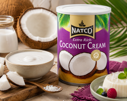 A can of NATCO Extra Rich Coconut Cream is shown in a styled kitchen-like setting with bowls of cream, sliced coconuts, a wooden spoon with coconut flakes, and a small dessert on a plate. The can features a purple label with green palm leaves and white coconut imagery.
