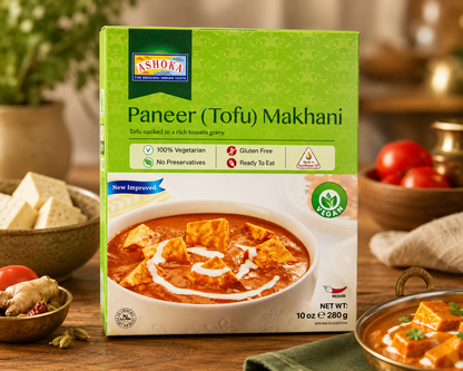 Ashoka Paneer (Tofu) Makhani ready meal box displayed on a wooden kitchen countertop with a bowl of creamy tomato tofu curry, fresh paneer cubes, tomatoes, ginger, and spices in a warm, softly blurred kitchen background.
