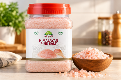 Aryan Himalayan Pink Salt 1kg jar displayed on a kitchen countertop beside a wooden bowl filled with coarse pink salt crystals, with scattered salt granules and a softly blurred kitchen background, ideal for showcasing pink salt for everyday cooking and seasoning.