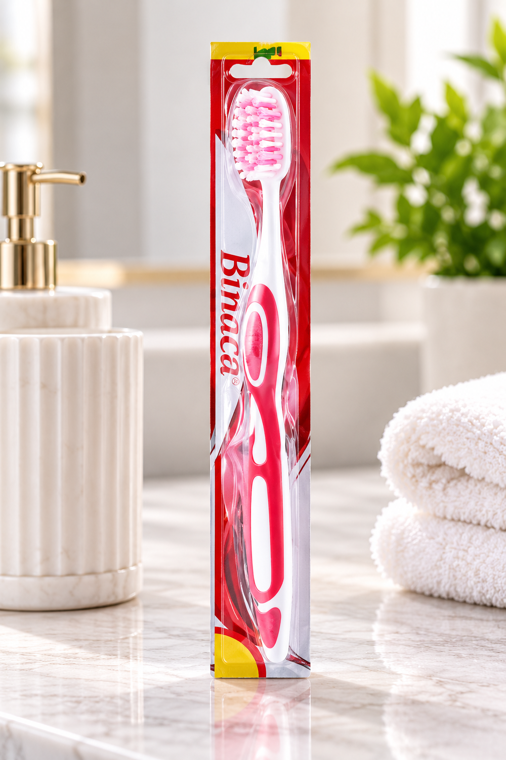 Binaca Soft Extra Clean Toothbrush in red and white packaging displayed upright on a glossy bathroom countertop, with a white ribbed holder, gold-toned dispenser, folded white towels, and green foliage in the background.