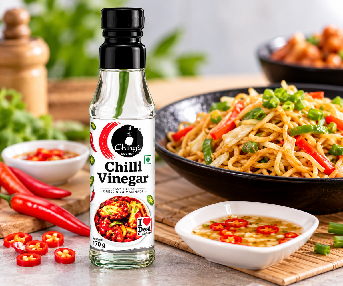 Ching’s Secret Chilli Vinegar 170g bottle displayed beside stir-fried noodles, a bowl of chilli dipping sauce and fresh red chillies on a kitchen table.
