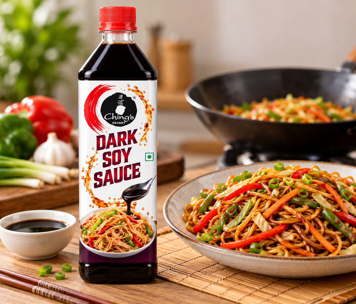 Ching’s Secret Dark Soy Sauce 750g bottle displayed beside a plate of stir-fried noodles with colourful vegetables, a bowl of soy sauce, fresh peppers and chopsticks on a bamboo kitchen mat.