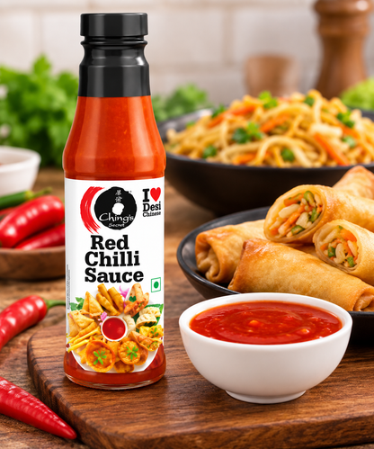 Ching’s Secret Red Chilli Sauce bottle displayed beside a bowl of red chilli sauce, crispy vegetable spring rolls, stir-fried noodles, fresh red chillies and green herbs on a wooden kitchen board.