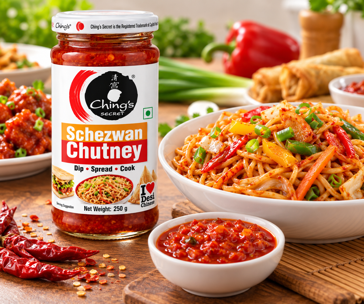 Ching’s Secret Schezwan Chutney 250g jar displayed beside a bowl of stir-fried noodles with colorful vegetables, a small bowl of red Schezwan chutney, crispy spring rolls, and dried red chillies on a wooden tabletop with fresh vegetables in the background.