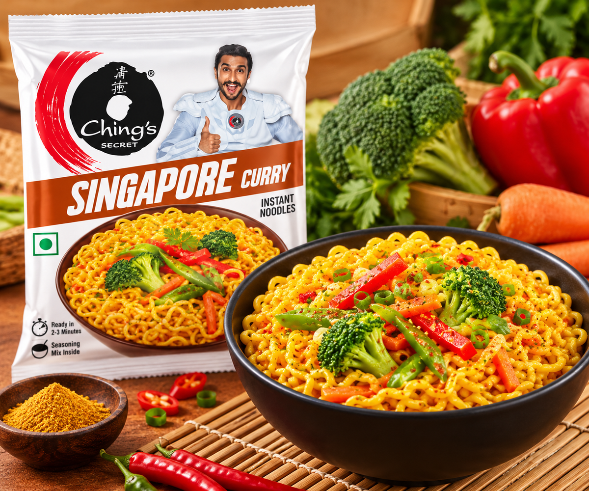 Ching’s Secret Singapore Curry Instant Noodles 60g packet displayed beside a black bowl of freshly prepared golden curry noodles with broccoli florets, red and green bell peppers, carrots, and fresh herbs. The white packet features the Ching’s Secret logo, Singapore Curry branding, a bowl-of-noodles illustration, and a smiling man giving a thumbs-up. Fresh vegetables, red chillies, curry seasoning, and wooden chopsticks arranged on a warm wooden surface create an inviting food photography scene.