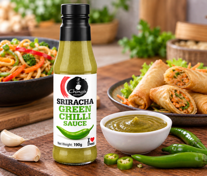 Ching’s Secret Sriracha Green Chilli Sauce 190g bottle displayed beside a bowl of green chilli sauce, fresh green chillies, garlic, vegetable noodles and crispy spring rolls on a wooden kitchen counter.