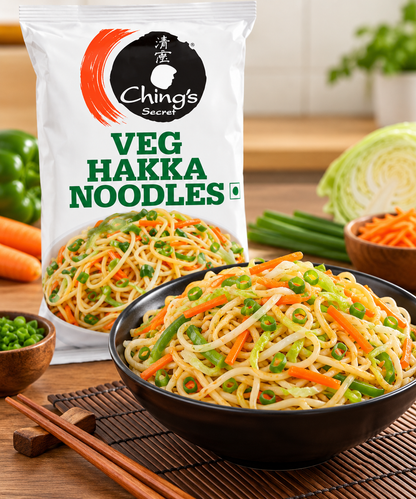 Ching’s Secret Veg Hakka Noodles packet displayed behind a black bowl of stir-fried Hakka noodles with carrots, green peppers, cabbage and spring onions, served on a wooden kitchen table with chopsticks and fresh vegetables.