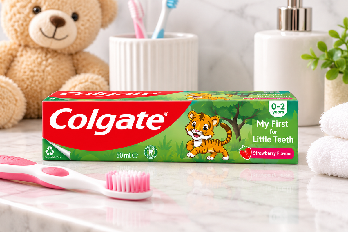 Colgate Junior Strawberry Flavour Toothpaste 50ml box displayed on a bright bathroom countertop beside a pink-and-white children's toothbrush, with a teddy bear, white towel, toothbrush holder, and green plant in the background.