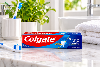Colgate Maximum Cavity Protection Regular Toothpaste 100ml box displayed on a marble bathroom countertop beside a blue and white toothbrush, toothpaste on the brush, a white ceramic toothbrush holder and folded white towel.