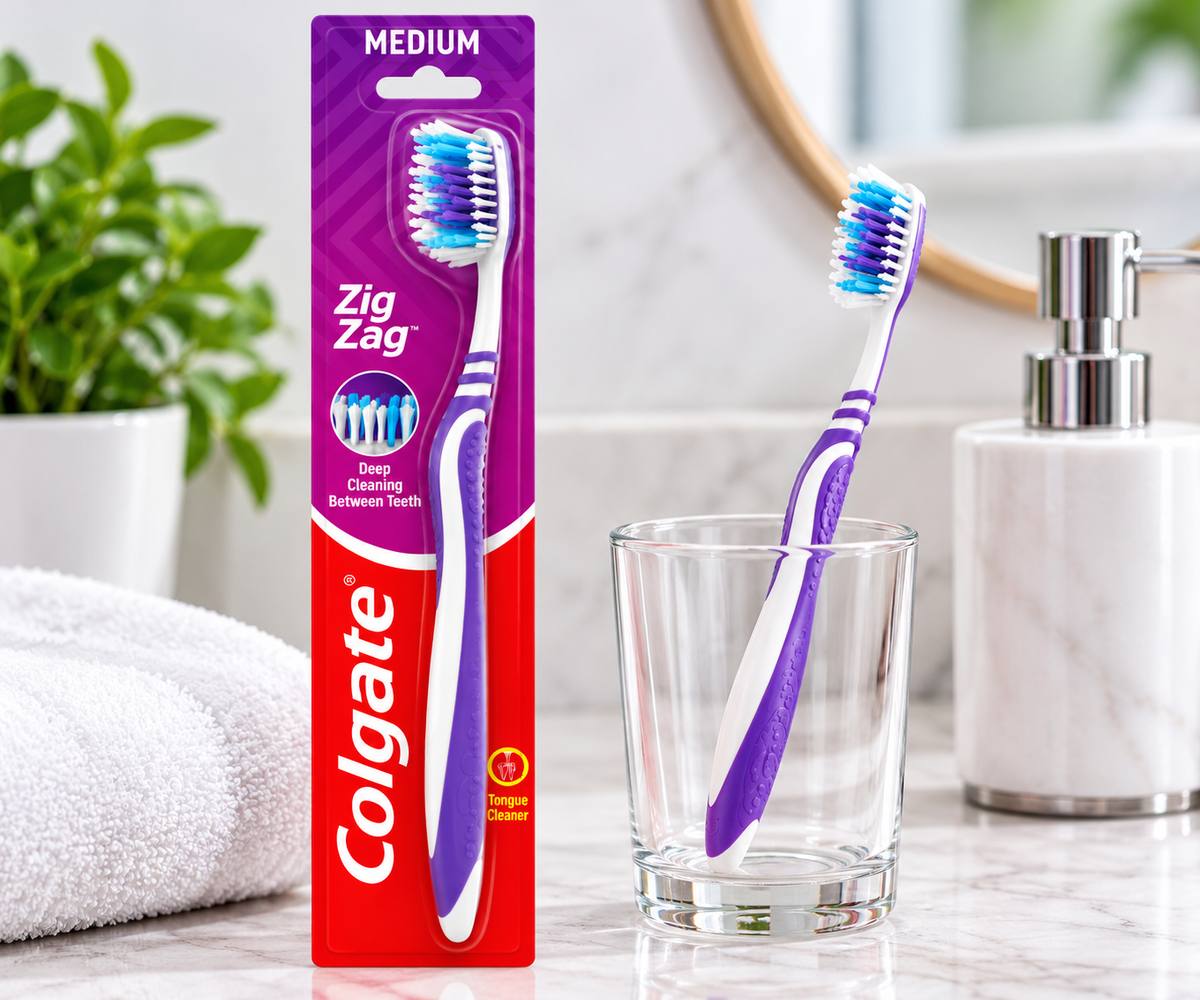 Colgate Zig Zag Toothbrush in purple and red packaging displayed on a bright bathroom countertop beside a white and purple toothbrush in a clear glass, with a folded white towel and green plant.