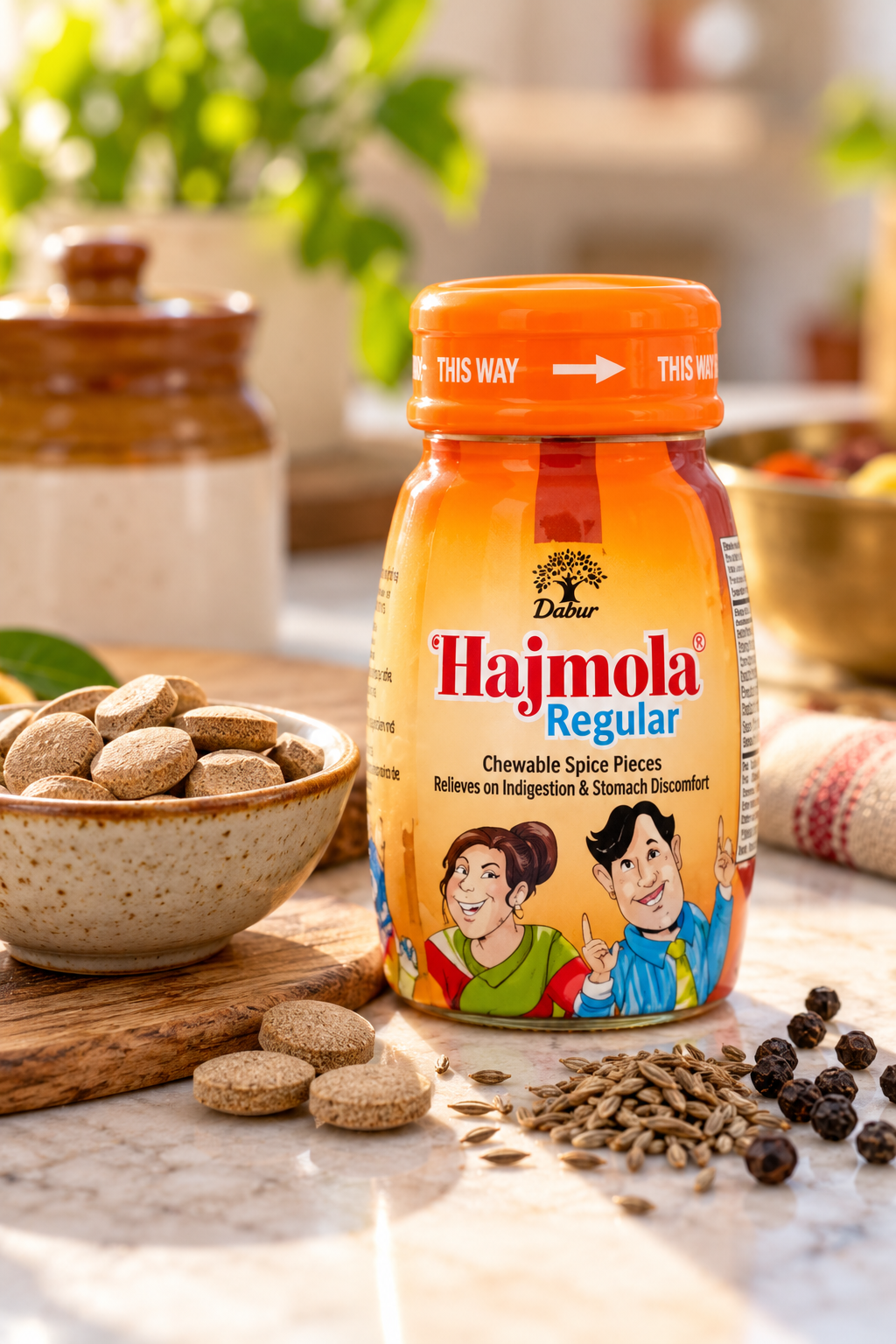 Dabur Hajmola Regular chewable spice tablets in an orange jar displayed on a kitchen countertop beside a small bowl of brown tablets and assorted whole spices in warm natural lighting.