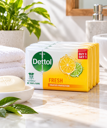 Dettol Fresh Bar Soap four-pack containing 4 × 100g bars displayed on a bright bathroom countertop beside a white soap bar, a folded towel, and green leaves, with lemon and lime illustrations on the yellow-orange packaging.