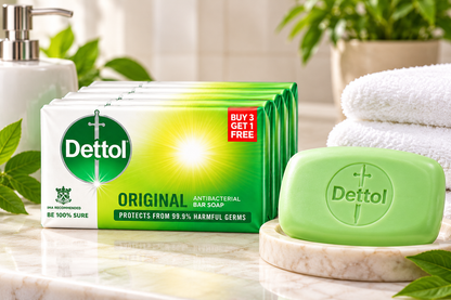 Dettol Original Bar Soap four-pack containing 4 × 100g bars displayed on a bright bathroom countertop beside a green Dettol soap bar, folded white towels, a silver dispenser, and green plants.