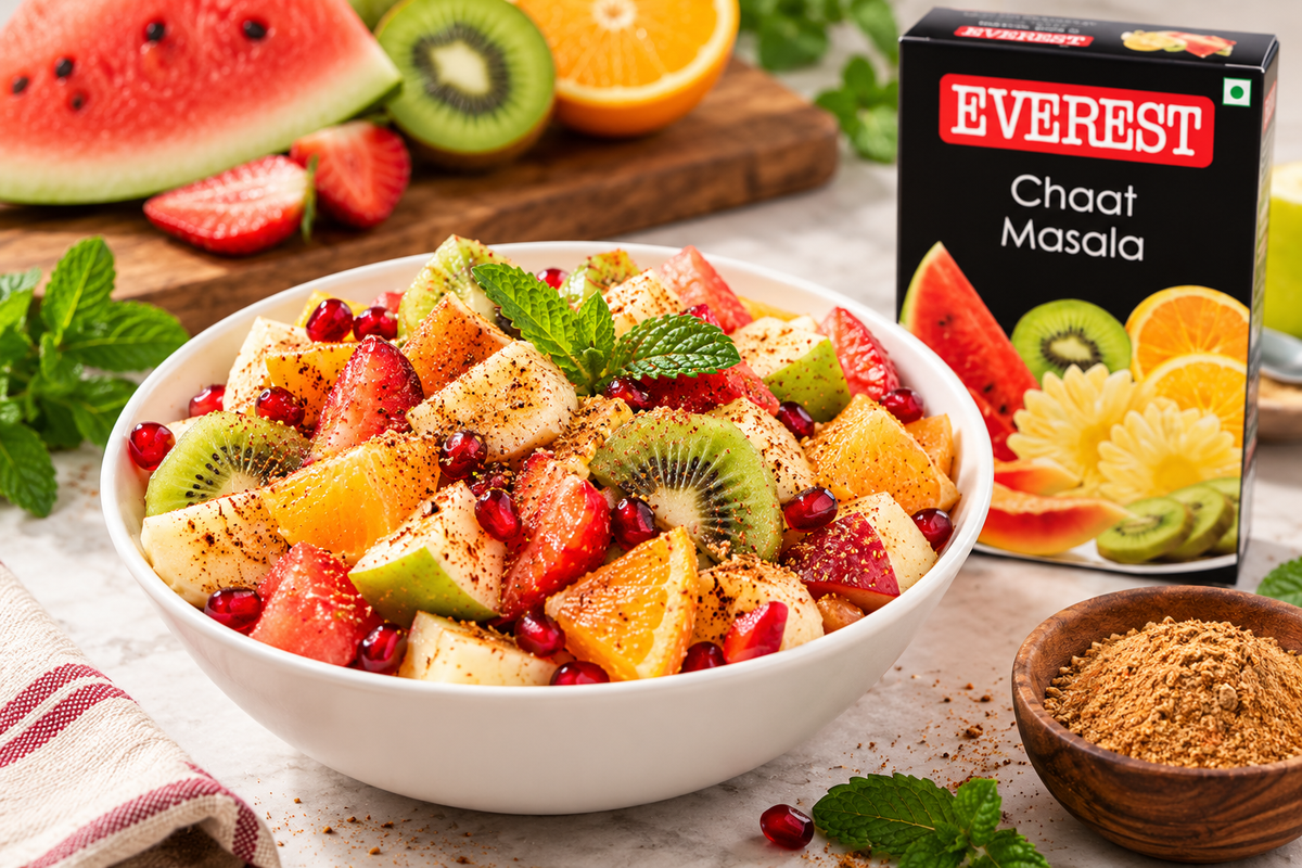 Everest Chaat Masala spice mix displayed beside a freshly prepared bowl of colorful fruit chaat made with watermelon, kiwi, orange, apple, strawberries, and pomegranate seeds, lightly seasoned with chaat masala and garnished with fresh mint leaves. The Everest Chaat Masala packet stands in the background alongside fresh fruit, highlighting the spice blend's use in preparing flavorful Indian fruit chaat and refreshing homemade snacks.