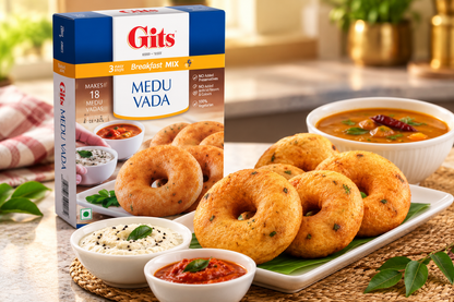 Gits Medu Vada Mix 500g box displayed beside golden, crispy Indian medu vadas served on a platter with coconut chutney, red chutney and a bowl of sambar on a warm kitchen counter.