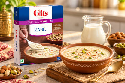Gits Rabdi Dessert Mix 100g box displayed beside a bowl of creamy Indian rabdi garnished with almonds and pistachios, with a glass jug of milk and assorted nuts on a warm kitchen counter.