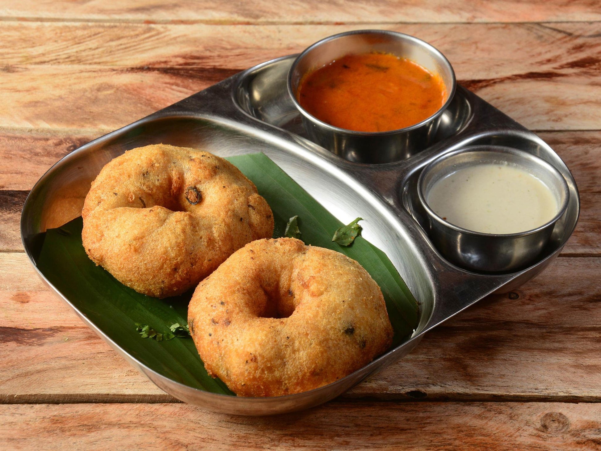Two golden brown medu vadas served on a banana leaf in a stainless steel tray with bowls of sambar and coconut chutney.
