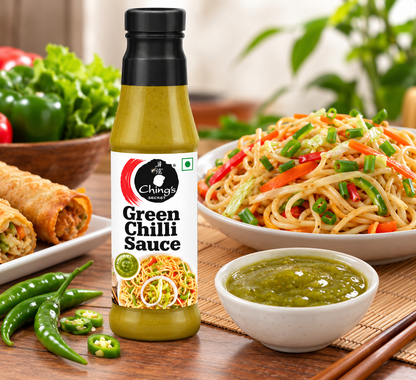Ching’s Secret Green Chilli Sauce bottle displayed beside a bowl of green chilli sauce and vegetable Hakka noodles, with fresh green chillies, spring onions and garlic on a wooden tabletop.