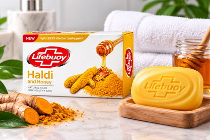 Lifebuoy Haldi and Honey Soap 123g displayed beside a yellow Lifebuoy soap bar on a wooden soap dish, with fresh turmeric roots, turmeric powder, honey and green leaves on a marble bathroom counter.