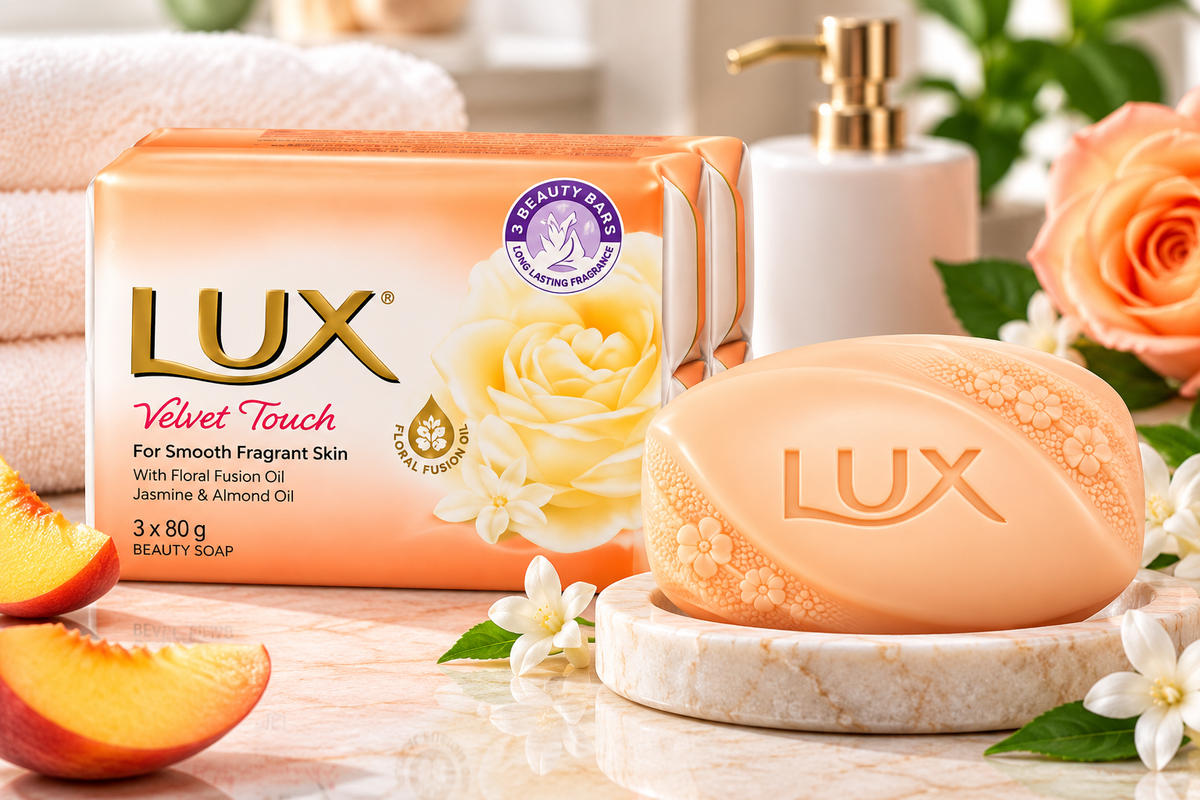 LUX Velvet Touch Soap 3-pack of three 80g bars displayed beside a peach-colored soap bar on a marble dish, with cream flowers, peach slices, green leaves, and folded white towels in a softly lit bathroom setting.