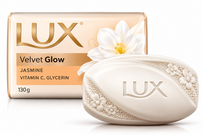 Lux Velvet Glow Jasmine Soap 130g featuring a smooth ivory oval soap bar embossed with the Lux logo and delicate floral detailing, paired with cream and gold packaging displaying the Lux branding, Velvet Glow name, and white jasmine flower illustrations. Showcased against a clean white background, highlighting the jasmine-scented beauty soap with vitamin C and glycerin for a luxurious daily bathing experience.