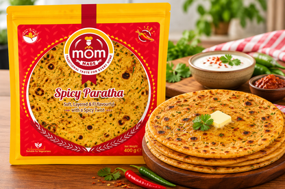 Mom Made Spicy Paratha 400g pack displayed beside a stack of golden, herb-speckled spicy parathas topped with butter and fresh coriander, served with yogurt and red chutney on a wooden table with fresh green and red chillies.