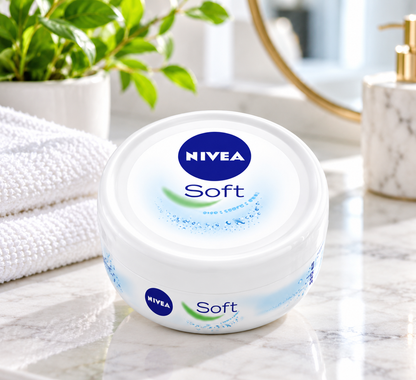 NIVEA Soft moisturizing cream pot displayed on a marble bathroom countertop beside a folded white towel, green plant and skincare accessories in a bright, modern bathroom setting.