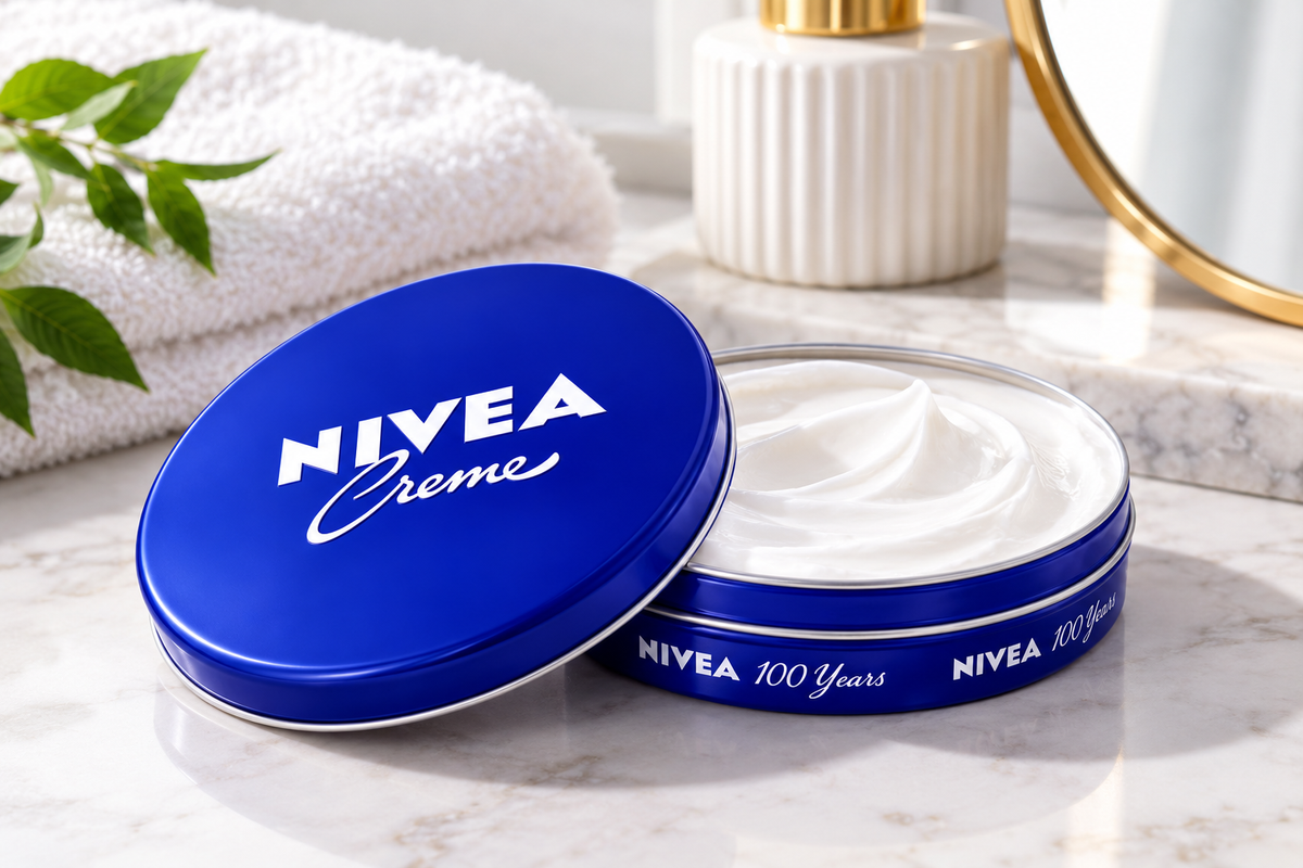 NIVEA Creme 60ml blue tin shown open with white cream inside, its blue lid resting beside it on a marble bathroom countertop with a folded white towel, green foliage, and a white dispenser in a bright, spa-inspired setting.