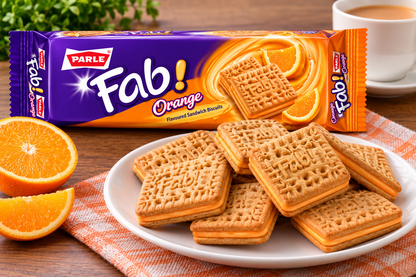 Parle Fab Orange Cream Biscuits packet displayed behind a plate of golden sandwich biscuits with orange cream filling, fresh orange slices and a cup of tea on a wooden table.