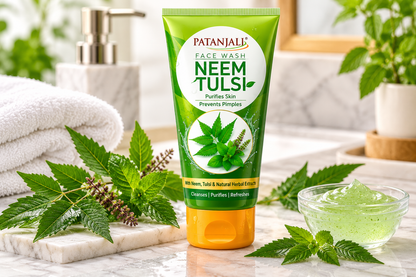 Patanjali Neem Tulsi Face Wash 600g green tube with an orange flip-top cap, white oval label, Patanjali branding, and neem and tulsi leaf illustrations displayed upright on a marble bathroom countertop. Surrounded by fresh neem leaves, tulsi sprigs, a clear glass bowl of green gel, and a soft white towel, with a leafy plant and modern bathroom accessories in the background. This bright, nature-inspired lifestyle image highlights the Ayurvedic herbal face wash as part of a refreshing daily facial cleansing r