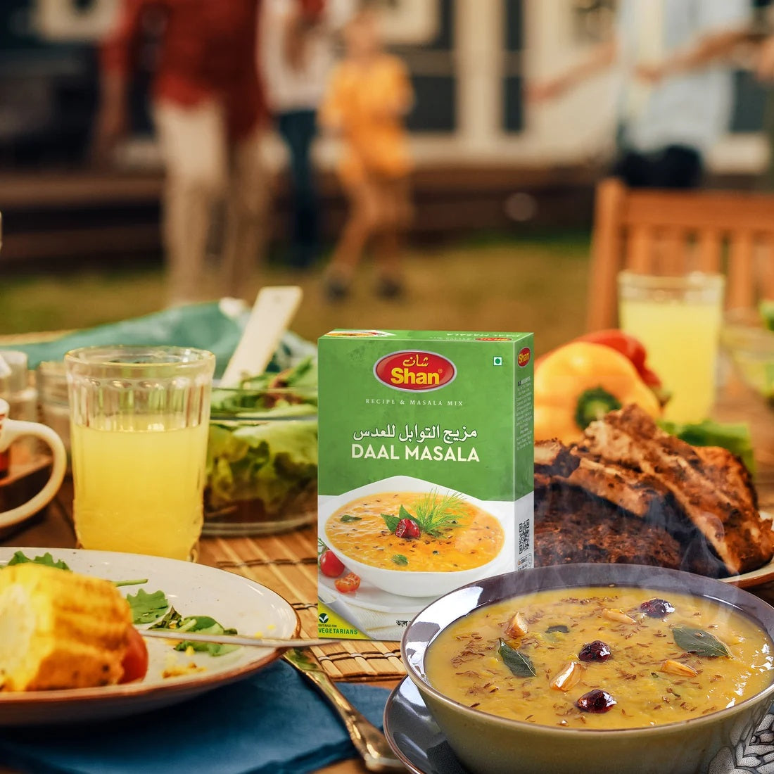 A box of Jhpie Shan Daal Masala seasoning mix placed on a dining table surrounded by a bowl of prepared dal, bread, drinks, and an outdoor patio background with blurred people.